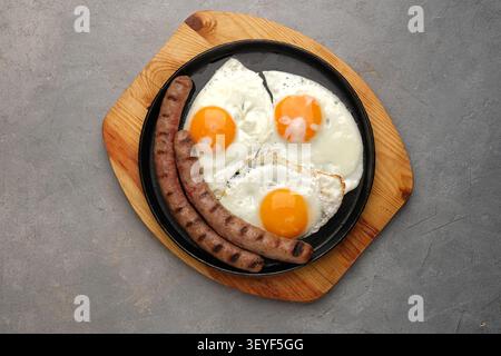 Gegrillte Würstchen mit Spiegeleiern, serviert in schwarzer Pfanne auf Holzbrett über grauer Steinoberfläche Stockfoto