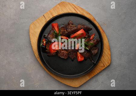 Gegrillte Rindfleischstücke mit Tomaten und Kräutern, serviert auf schwarzem Teller über Holzbrett Stockfoto