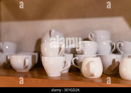 Zahlreiche Kaffeetassen und Tassen aus weißer Keramik sind übersichtlich angeordnet und auf einem Holzregal gestapelt. Das Bild weist eine geringe Schärfentiefe mit warmem A auf Stockfoto