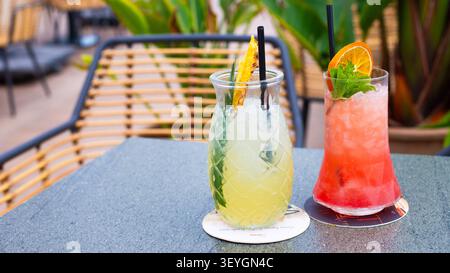 Zwei farbenfrohe Cocktails auf einem Tisch im Freien, einer mit einer Ananusscheibe und der andere garniert mit einer Orangenscheibe und Minzblättern Stockfoto
