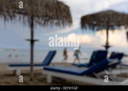 Unscharfe Strandresort-Szene mit Strohschirmen, blauen Liegestühlen und Menschen, die bei Sonnenuntergang am Meer spazieren gehen. Perfekter Hintergrund für einen Sommerurlaub Stockfoto