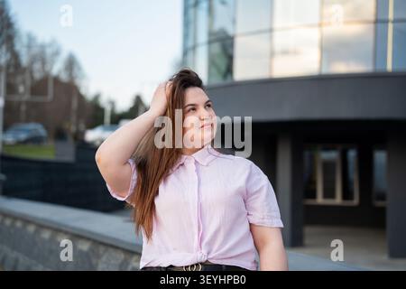 Selbstbewusste junge Berufsfrau in lässiger Kleidung vor dem modernen Glasbürogebäude. Corporate Lifestyle städtischer Arbeitsplatz und Karriereerfolg. Stockfoto