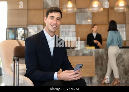 Ein lächelnder Mann sitzt in der Hotellobby mit Koffer und Smartphone Stockfoto