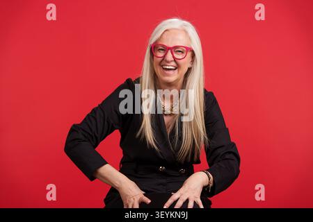 Seniorin lacht mit rosa Brille auf rotem Hintergrund Stockfoto