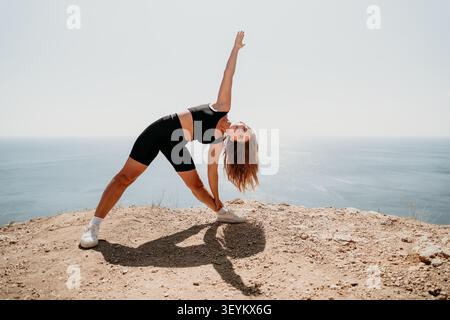 Yoga Stretching an den Klippen junge Frau in schwarzer Sportbekleidung, die an einem sonnigen Tag mit Blick auf den Ozean eine Dreieckspose für Wellness übt Stockfoto