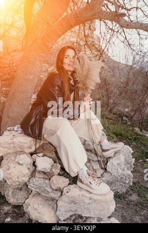 Frau Natur Pampas Gras lächelnde junge Frau sitzt auf Felsen an einem Baum mit getrocknetem Schilf unter warmem Sonnenlicht Stockfoto