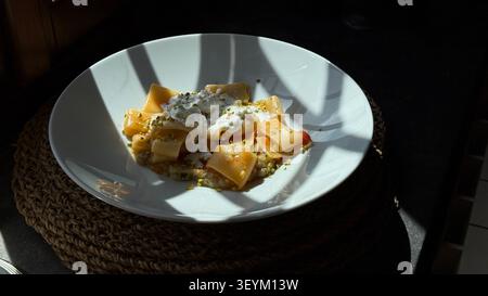 Köstliche Gourmet-Pasta mit cremiger Sauce und Pistazien, serviert in einer eleganten weißen Schüssel. Die Platte wird künstlerisch in verblühtem Sonnenlicht platziert Stockfoto