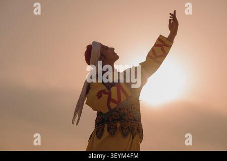 Die Volkstänzerin in aufgestickter traditioneller Tracht hebt den Arm triumphierend gegen den glühenden, goldenen Wüstenhimmel mit dramatischem, hintergrundbeleuchtetem Sonnenschein und Stockfoto