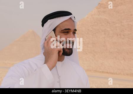 Fröhlicher arabischer Profi in traditionellem Keffiyeh und du lachst mitten im Handygespräch draußen mit Pyramiden als Bokeh-Hintergrund Stockfoto