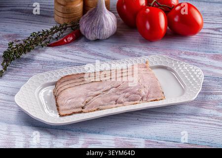 Pastrami in Scheiben auf weißem, strukturiertem Teller mit frischen roten Tomaten, Knoblauchzweig Chili-Pfeffer auf rustikaler Holzoberfläche Stockfoto