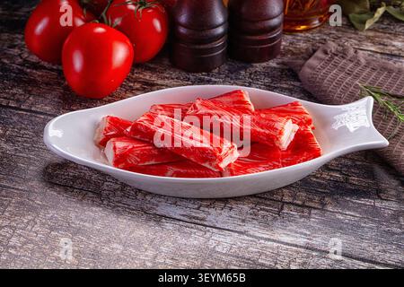 Leuchtend rote Krabbenstangen in einer weißen Keramikschale in Form von Fischen. Frische Tomaten dunkle Gewürzmühlen aus Holz im Hintergrund. Rustikale Holzoberfläche mit Stockfoto