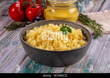 Couscous serviert in dunkler Keramikschale, garniert mit Petersilie. Rote Tomaten, Rosmarin, Glas mit trockenem Couscous im Hintergrund. Rustikale Holzfläche mit Stockfoto