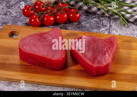 Zwei rohe Thunfischsteaks auf Holzbrett mit Rosmarin aus Kirschtomaten. Rustikale Küche mit grauem Steinhintergrund. Warmes Licht hebt satte Re hervor Stockfoto