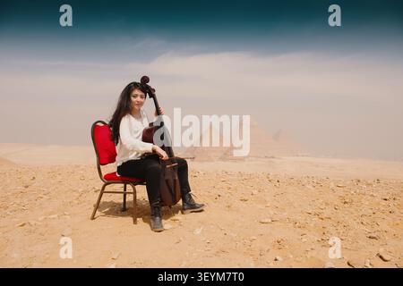 Junge Musikerin, die Kontrabass auf rotem Stuhl in der ägyptischen Sandwüste spielt, mit den ikonischen Pyramiden von Gizeh im Hintergrund, ideal für klassische Musikkunst Stockfoto