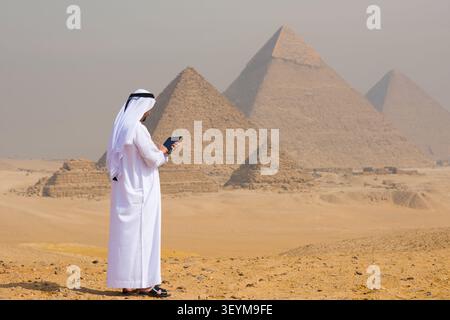 Arabischer Mann mit traditionellem weißem Thobe und Keffiyeh, der ein digitales Tablet verwendet, während er auf dem Gizeh-Plateau steht, auf dem sich die legendären Pyramiden durch die Wüste erheben Stockfoto