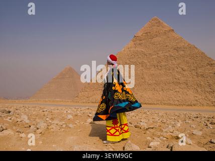 Eine kunstvolle Figur mit traditioneller Tracht steht vor den legendären Pyramiden von Gizeh auf dem ägyptischen Sandplateau, einer zeitlosen Kulturreiseszene Stockfoto