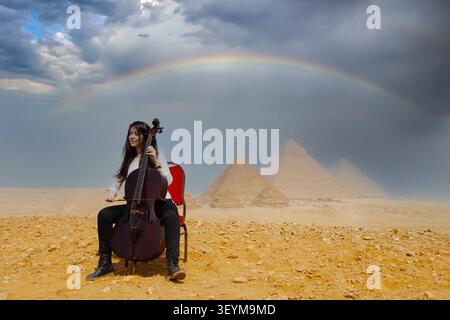 Junge Cellistin, die klassische Musik im Freien vor den Großen Pyramiden von Gizeh unter einem Regenbogen und stürmischen dramatischen Himmel in Ägypten vorführt Stockfoto