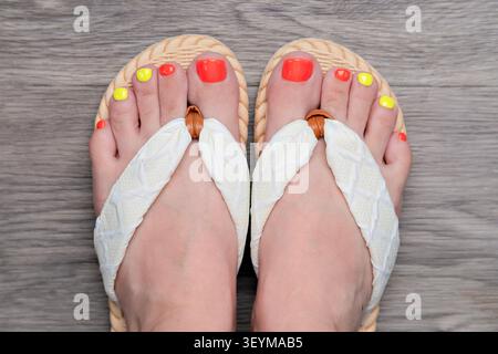 Blick von oben auf Frauenfüße in weißen Flipflops mit farbenfroher Neon-Pediküre auf Holzboden. Stockfoto