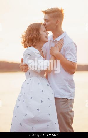 Ein junger Mann küsst seiner lächelnden Freundin auf die Stirn, während sie sich während der goldenen Stunde am Wasser umarmen und eine romantische und freudige Verbindung einfangen Stockfoto