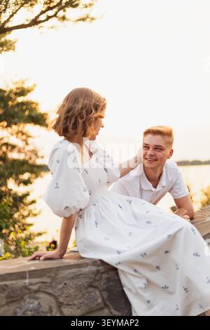 Ein glückliches junges Paar teilt einen zärtlichen Blick, während es während der goldenen Stunde auf einer Steinmauer sitzt und auf ein Gewässer blickt Stockfoto