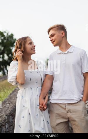 Ein lächelndes junges Paar in lässiger Sommerkleidung geht Hand in Hand auf einem Steinweg und genießt einen warmen gemeinsamen Tag Stockfoto