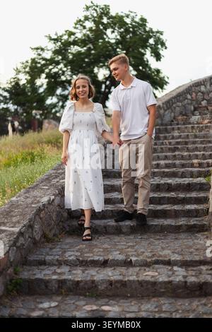Ein glückliches junges Paar geht Hand in Hand eine verwitterte Steintreppe hinunter, die von Grün und einem großen Baum umgeben ist Stockfoto