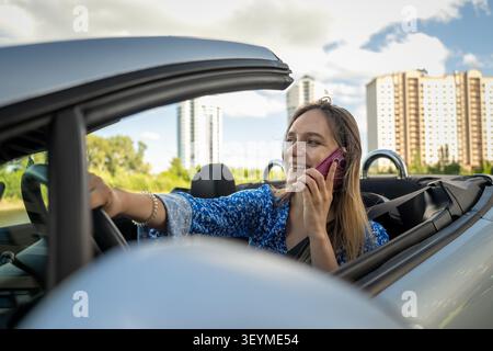 Lächelnde Frau telefoniert mit einem Smartphone, während sie in einem Cabrio sitzt. Moderne Kommunikation und glücklicher urbaner Lebensstil. das Konzept von unsicherem Fahren Stockfoto