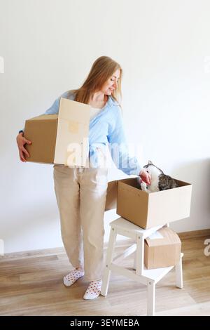 Umzugstag mit einem geliebten Haustier. Die junge Frau organisiert Boxen, während ihre neugierige Katze das neue Zuhause erforscht. Stockfoto