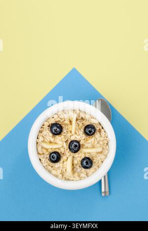 Weißes Gericht mit Hafer, Bananen und Heidelbeeren auf blauem und gelbem Hintergrund Stockfoto