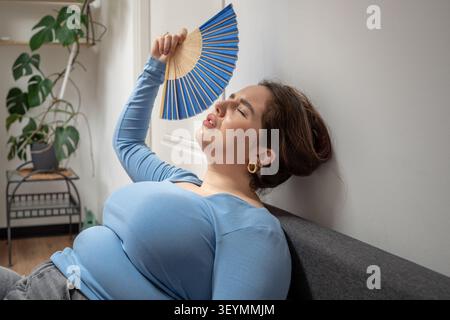 Erschöpfte, übergewichtige junge Frau auf Sofa, die unter Überhitzung im Innenbereich leidet, indem sie einen Handventilator benutzt, um sich abzukühlen, und schwer atmet. Verstopfte Luft, Unwohlsein, Mangel Stockfoto