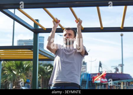 Sportlicher bärtiger Mann, der an der horizontalen Außenbar im Seaside Park zieht, männliche Athleten trainieren Oberkörperkraft auf Straßentrainingsgeräten, Konzept der Fitness Ausdauer und aktiver Lebensstil Stockfoto