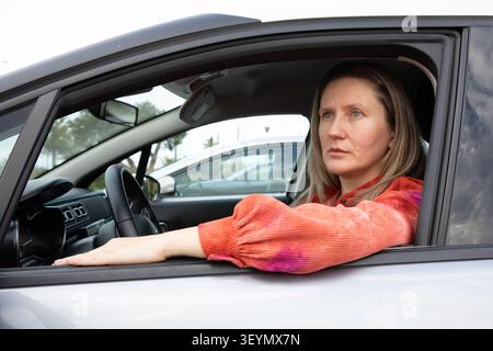 Fahrerin mit blonden Haaren sitzt im Auto auf dem Parkplatz. Sie blickt mit einem fokussierten Ausdruck nach draußen. Horizontal. Nahaufnahme. Stockfoto