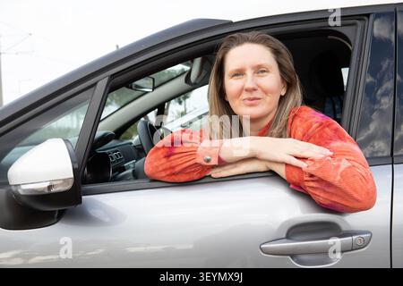 Fahrerin mit blonden Haaren sitzt im Auto mit Armen vor dem Fenster. Sie lächelt, während sie tagsüber in der Stadt parkt. Horizontal. Stockfoto