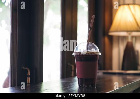 Eiskaffee in klarem Glas mit Abdeckung auf Holztisch an den Fenstern und Lichter im Hintergrund Stockfoto