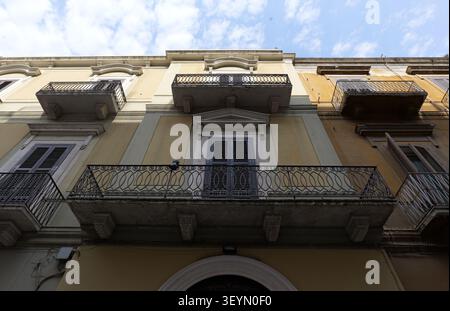 Fassade des Gebäudes mit kunstvoll verzierten schmiedeeisernen Balkonen, geschlossenen Fenstern mit Holzläden und einer gelben, hellen Wand unter blauem, bewölktem Himmel, Showcasi Stockfoto