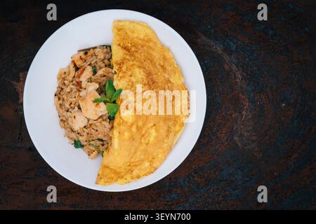 Hausgemachter thailändischer würziger gebratener Reis mit gebratenem Huhn und Basilikum bedeckt mit einem halbkreisförmigen Omelett auf weißem Teller mit Löffel in weißer Serviette auf Holz Stockfoto