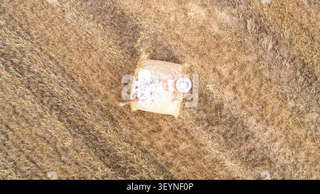 Luftbild aus der Vogelperspektive einer modischen Frau, die auf einem großen runden Heuballen in einem abgeernteten Feld mit goldenem Getreide liegt. Sie hält einen Hut mit breiter Krempe. Stockfoto