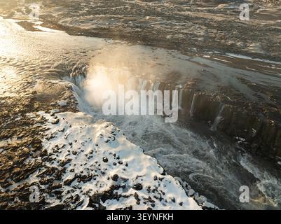 Aus der Vogelperspektive des Dettifoss-Wasserfalls mit Nebel, der von der mächtigen Kaskade über zerklüftete vulkanische Felsen und schneebedecktes Gelände in Nordurthing, Icel, aufsteigt Stockfoto