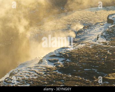 Aus der Vogelperspektive des Dettifoss-Wasserfalls mit Nebel, der von der mächtigen Kaskade über zerklüftete vulkanische Felsen und schneebedecktes Gelände in Nordurthing, Icel, aufsteigt Stockfoto
