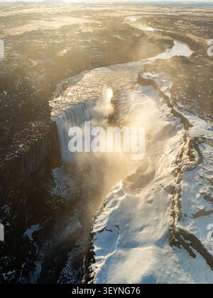 Blick aus der Vogelperspektive auf den Dettifoss-Wasserfall, umgeben von schneebedecktem vulkanischem Gelände und Nebel unter der goldenen Stunde in Nordurthing, Island. Stockfoto