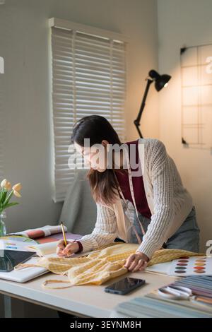 Asiatische Modedesignerin zeichnet in kreativer Form Kleidungsskizzen mit Stoffmustern und Farbpaletten auf dem Tisch Stockfoto