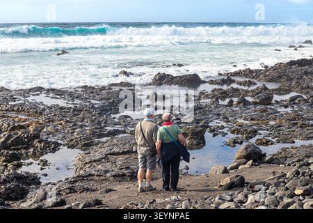 Ein paar Touristen mittleren Alters, die die schwere See genießen, die auf den Vulkanfelsen von El Golfo an der Südwestküste von Lanzarote bricht Stockfoto