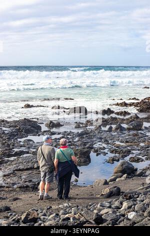 Ein paar Touristen mittleren Alters, die die schwere See genießen, die auf den Vulkanfelsen von El Golfo an der Südwestküste von Lanzarote bricht Stockfoto