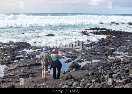 Ein paar Touristen mittleren Alters, die die schwere See genießen, die auf den Vulkanfelsen von El Golfo an der Südwestküste von Lanzarote bricht Stockfoto