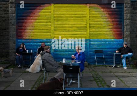 Menschen sitzen an Cafétischen mit ihren Hunden vor einem farbenfrohen, hell bemalten Rolltor, das einen regenbogenartigen Sonnenaufgang darstellt. Stockfoto
