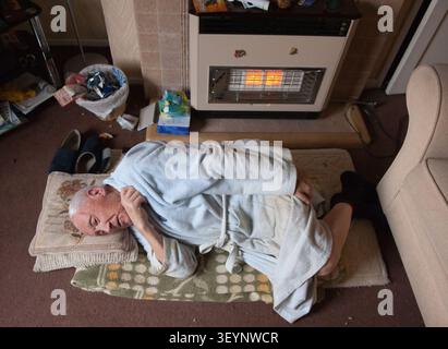 Ein älterer Mann in einem Bademantel liegt auf einem provisorischen Bett auf dem Boden neben einem Gasheizofen, was Kraftstoffarmut und Sozialfürsorge hervorhebt. Stockfoto