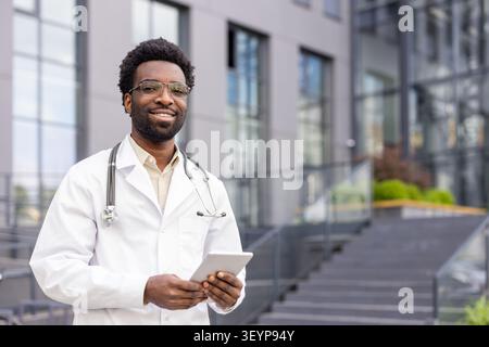 Lächelnder männlicher Arzt mit weißem Mantel und Stethoskop, hält ein digitales Tablet, während er selbstbewusst vor einer modernen Gesundheitseinrichtung steht Stockfoto