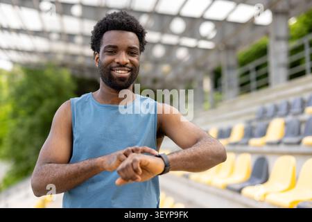 Ein lächelnder Athlet fühlt sich selbstbewusst und überwacht den Fortschritt seiner Smartwatch, verfolgt Leistungs- und Wellnessziele während eines Fitnesstrainings in einem Sportstadion im Freien Stockfoto