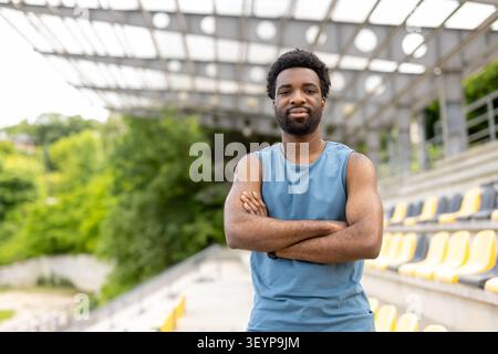 Selbstbewusster Athlet posiert mit gefalteten Armen, blickt direkt in die Kamera, trägt ein ärmelloses Hemd, steht bereit für Sport oder Training in einem leeren Stadion mit gelben und grauen Sitzen Stockfoto