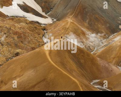 Aus der Vogelperspektive auf Kerlingarfjoll mit zwei Personen, die auf einem Kamm stehen, umgeben von dampfenden geothermischen Lüftungsöffnungen, Schneestücken und lebhaften orangen vulkanischen m Stockfoto